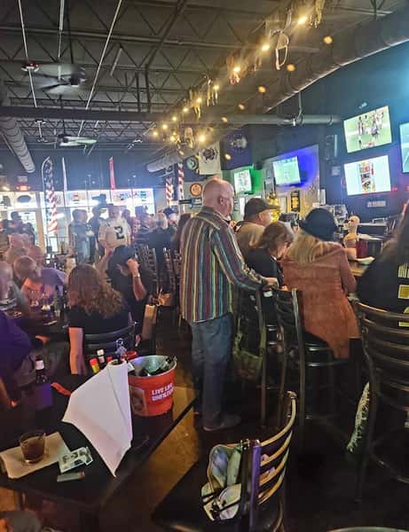 Busy sports bar scene with crowded bar seating, patrons watching multiple TV screens, servers pouring beers, and a lively American pub atmosphere with warm string lighting and casual dining tables.