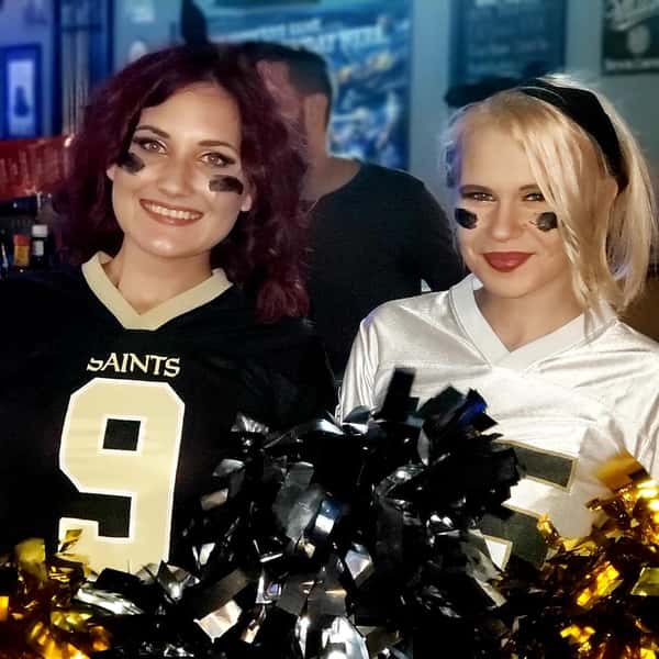 Two women in New Orleans Saints jerseys with face paint and pom-poms, smiling at a sports bar during game day, creating a fun and energetic football fan atmosphere.
