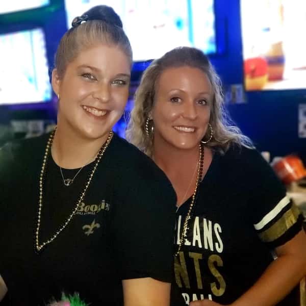 Two women smiling at a sports bar wearing New Orleans Saints shirts and beads, with TV screens in the background and a lively game-day atmosphere at Boogies Grill & Chill.