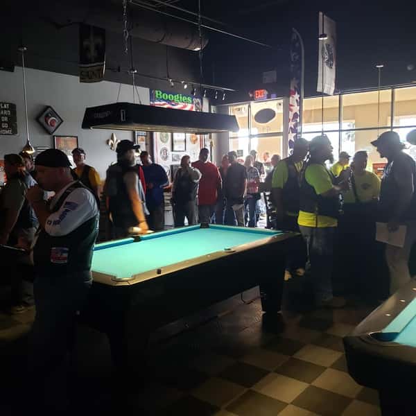 Crowded sports bar with patrons gathered around illuminated pool tables, casual drinks in hand, and a lively game-day atmosphere at Boogies Grill & Chill.
