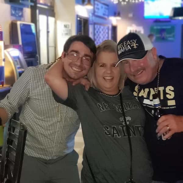 Three friends smiling and posing at a sports bar in New Orleans Saints apparel, holding drinks in a fun, social game-day environment with TVs and ambient lighting.