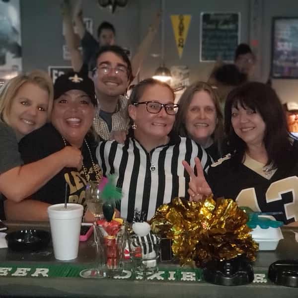 Group of friends at a lively sports bar smiling and posing at the counter, wearing New Orleans Saints gear, with festive drinks, shot glasses, and a fun game-day atmosphere.