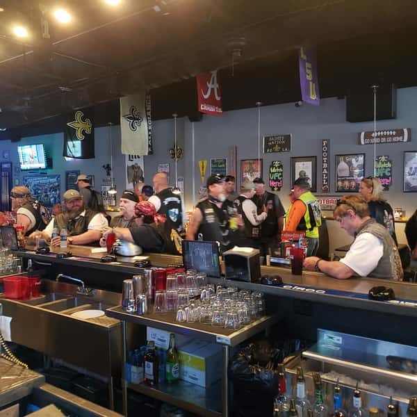 Busy bar counter at a sports pub with patrons in biker vests and Saints gear enjoying drinks, glassware lined up, and a high-energy American bar atmosphere.