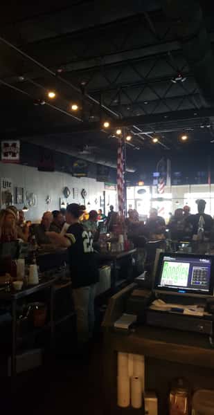 Busy sports bar interior with patrons gathered around a lively bar counter, warm ambient lighting, draft beers being served, and casual American pub atmosphere with TV screens and bar seating visible.