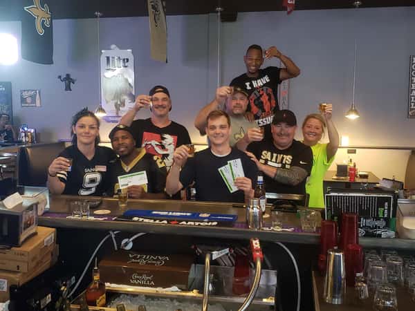 Group of sports bar staff and patrons raising shot glasses in celebration, wearing New Orleans Saints apparel, with bar counter, beer bottles, and lively game-day atmosphere in a casual American pub.