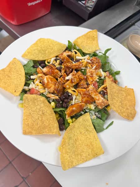 Fresh Southwest BBQ chicken salad topped with shredded cheese, black beans, corn, diced tomatoes, and tangy barbecue-glazed chicken over a bed of leafy greens, garnished with crispy tortilla chips on a white plate.