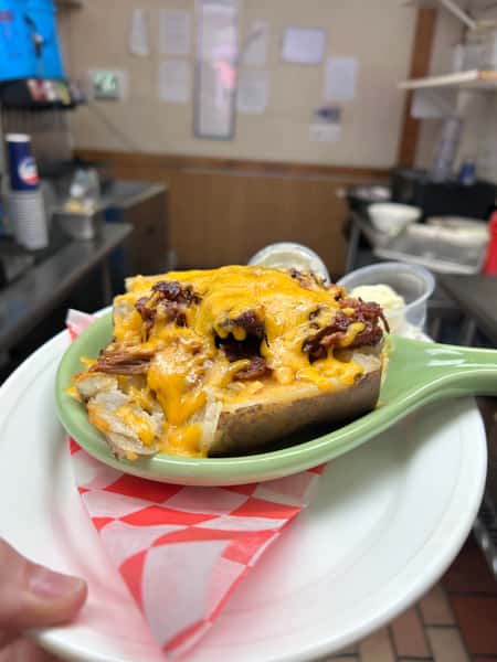 Baked potato loaded with pulled pork and melted cheddar cheese, served with sides of sour cream and butter on a green ceramic dish with checkered liner.