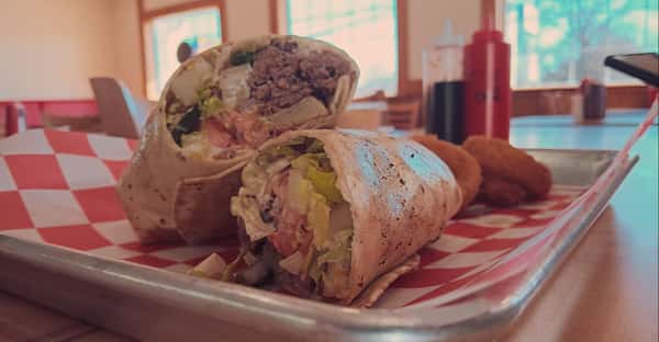 Grilled beef and veggie wrap cut in half, revealing lettuce, tomato, and ground beef, served with a side of hushpuppies on a checkered-lined tray in a casual dining setting.