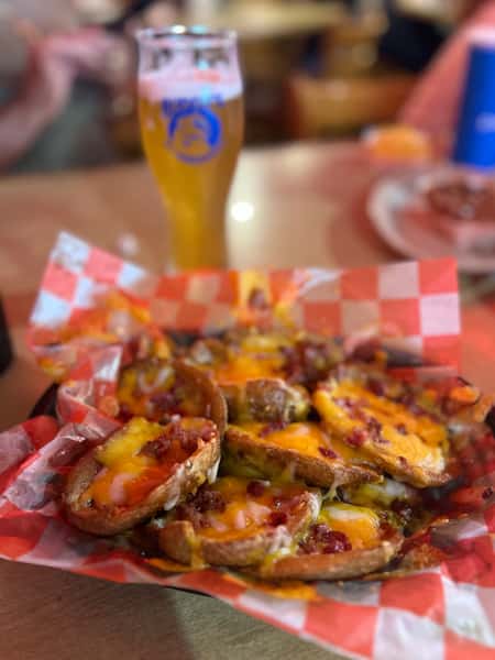 Crispy potato skins topped with melted cheddar cheese, bacon bits, and scallions, served on red checkered paper with a glass of draft beer in the background — perfect bar food.