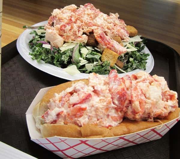 lobster roll and a caesar salad topped with lobster on a tray