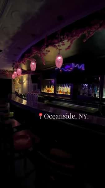 Stylish bar interior at Lushes in Oceanside, NY, with backlit liquor shelves, pink pendant lighting, and floral ceiling décor creating a trendy cocktail lounge atmosphere.