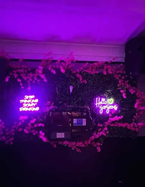 Instagram-worthy restaurant wall with pink cherry blossom décor and neon signs reading "Stop Thinking Start Drinking" and "Hello Gorgeous," set against lush greenery for a chic cocktail bar vibe.