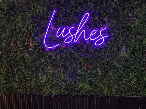 Purple neon "Lushes" sign mounted on a green plant wall, highlighting a stylish cocktail lounge with modern botanical décor and upscale nightlife ambiance.