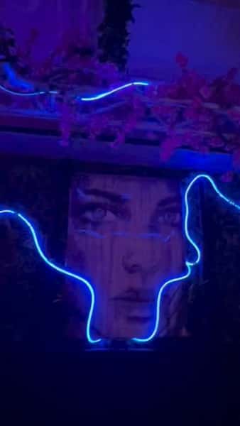Neon blue face outline framing a portrait mural inside a trendy lounge, with purple ambient lighting and floral décor creating a vibrant nightlife atmosphere.
