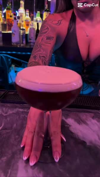 Bartender serving a frothy espresso martini in a coupe glass at an upscale New York cocktail bar, with illuminated liquor bottles and vibrant nightlife lighting in the background.