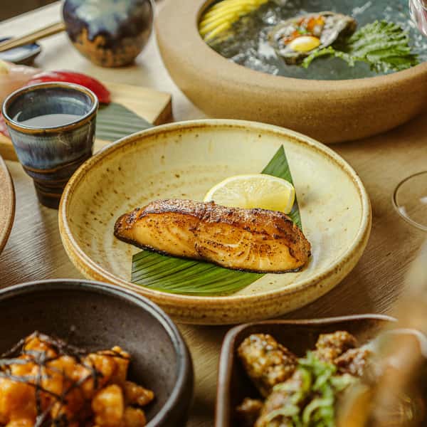 Miso-glazed salmon fillet on a bamboo leaf with a lemon wedge, surrounded by assorted Japanese dishes including sashimi and soy-glazed appetizers, on a wooden table.