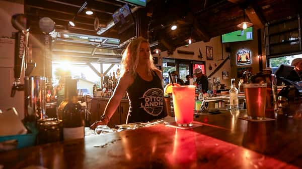 Bartender wearing an Otto's Landing tank top behind the bar at sunset, with cocktails and pints of beer glowing on the wooden counter inside the rustic, lively restaurant.