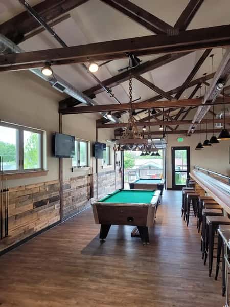 Rustic game room at Otto's Landing with exposed wood beams, two pool tables, mounted flat-screen TVs, and a long bar counter—ideal for casual drinks, games, and socializing in a laid-back atmosphere.