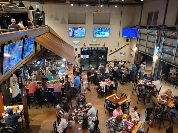 Overhead view of Otto's Landing's vibrant indoor dining area, showcasing a packed bar, flat-screen TVs streaming sports, and a bustling crowd enjoying food and drinks in a rustic, high-ceilinged space.