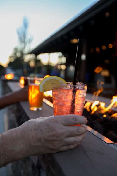 Hand holding a refreshing citrus cocktail with lemon garnish at Otto's Landing's fire pit patio, with flames and sunset in the background.
