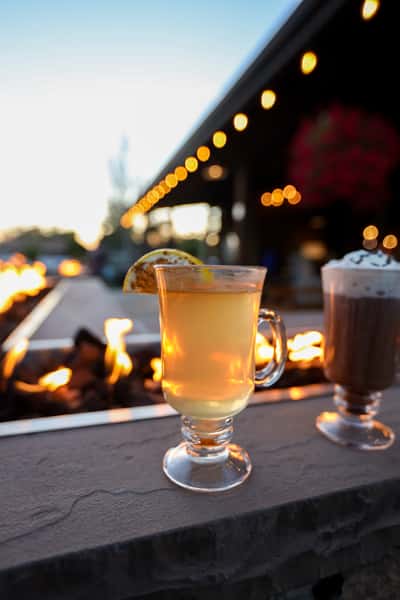 Hot toddy-style cocktail with a lemon wedge served in a glass mug beside Otto's Landing's fire pit, with string lights glowing in the background.