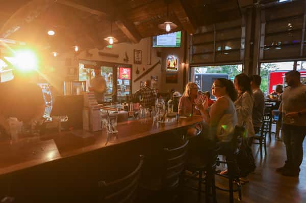 Warm sunset light illuminating patrons at a wooden bar inside a rustic restaurant, creating a cozy and inviting ambiance with casual seating and drinks in hand.