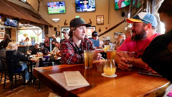 Two friends enjoying beers and cocktails at a high-top table inside Otto's Landing, with sports on TV and a lively dining crowd behind them.