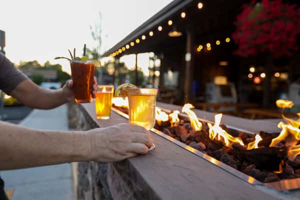 Guests holding a Bloody Mary and hot toddy-style cocktails near the outdoor fire pit at Otto's Landing, enjoying warm drinks at dusk.