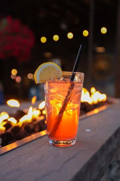 Bright citrus cocktail with lemon slice resting on Otto's Landing's fire pit ledge, glowing against string lights and evening ambiance.