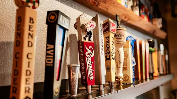 Close-up view of craft and domestic beer taps including Rainier, Coors Light, Modelo, and Bend Cider, displayed on a wood-accented bar wall inside a cozy taproom.