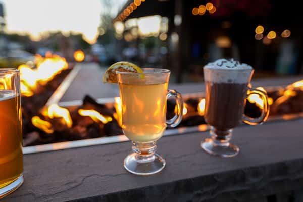 Hot toddy cocktail with lemon and a whipped cream-topped hot chocolate on Otto's Landing's outdoor fire pit ledge at dusk.