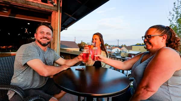 Smiling friends raising glasses of red beer on Otto's Landing's outdoor patio at sunset, enjoying a relaxed evening with rooftop views.