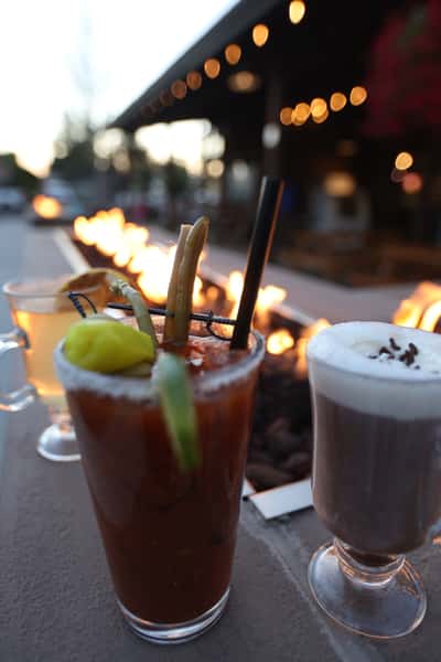 Bloody Mary cocktail and whipped-cream-topped hot cocoa placed beside Otto's Landing's outdoor fire pit during golden hour.