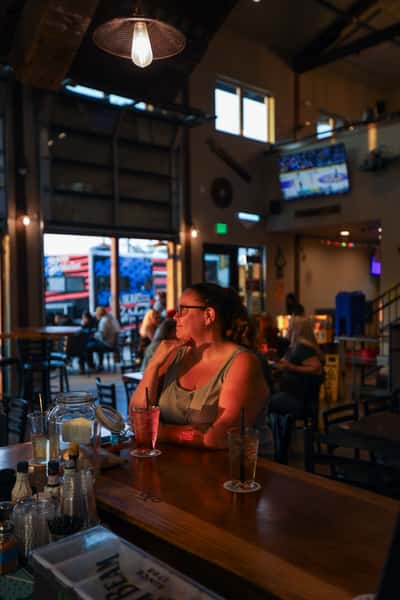 Woman enjoying a cocktail at the bar inside Otto's Landing, with views of the patio and evening light streaming through garage-style windows.