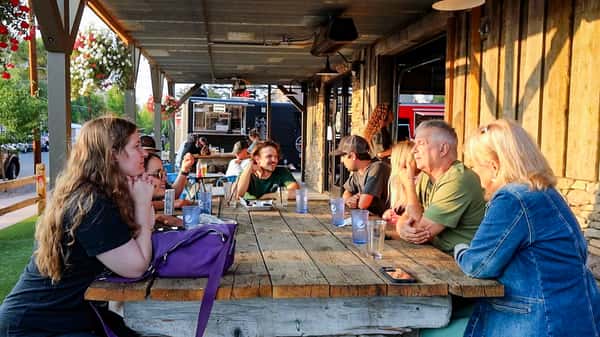 Groups of people dining and chatting at large wooden tables on a covered outdoor patio, surrounded by hanging flower baskets and rustic wooden siding.