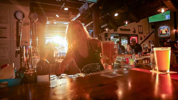 Group of friends enjoying pints of beer at a wooden table inside Otto's Landing, with sports on TV and a bustling bar scene in the background.