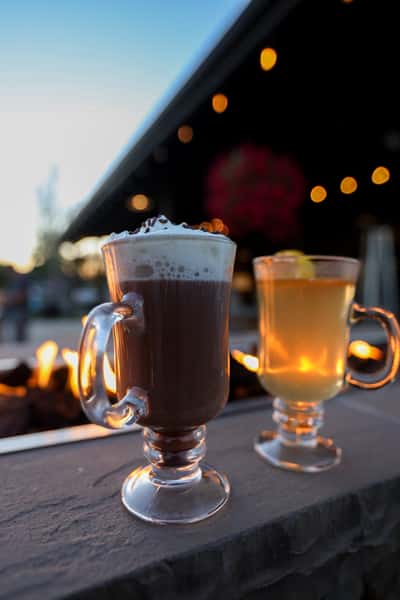 Close-up of a whipped-cream-topped hot chocolate and a hot toddy with lemon garnish set on the fire pit ledge at Otto's Landing, glowing in the warm evening light.