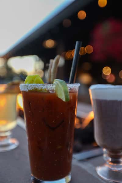 Close-up of a garnished Bloody Mary cocktail with lime wedges, celery, and salted rim at Otto's Landing, set against the glow of the outdoor fire pit.