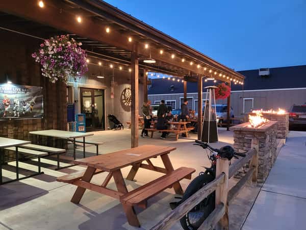 Cozy evening scene on Otto's Landing's outdoor patio, with picnic tables, hanging flower baskets, and glowing fire pits under string lights—perfect for casual dining and social gatherings.