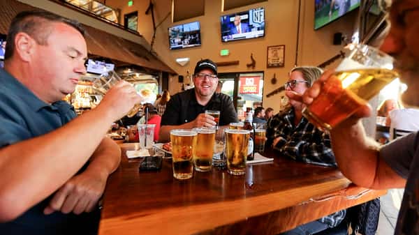 Group of friends enjoying pints of beer at a wooden table inside Otto's Landing, with sports on TV and a bustling bar scene in the background.