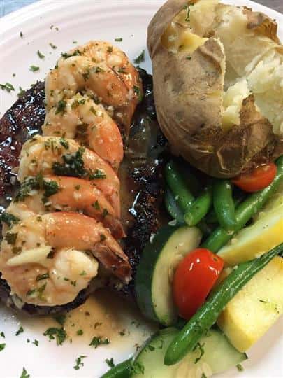 a steak with six shrimp and a side of baked potato with zucchini, tomato and green beans