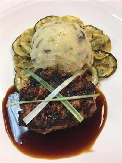 a steak in a pool of gravy with a side of zucchini and mashed potato