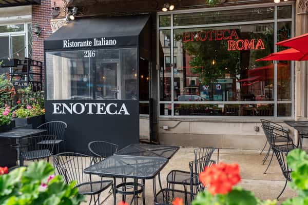 Charming outdoor patio dining area with black wrought-iron tables, vibrant flower planters, and red umbrellas lining the sidewalk — perfect for al fresco meals outside Letizia's Natural bakery and café.