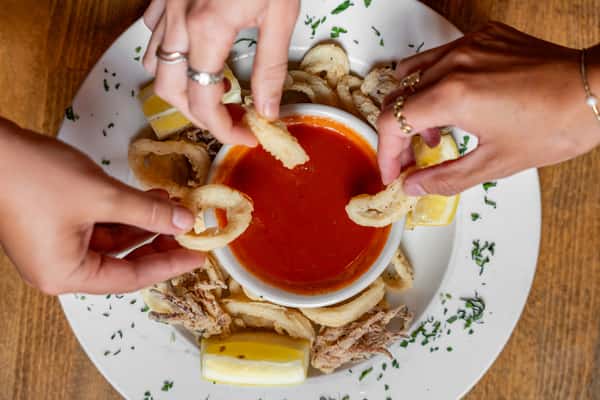 Hands reaching for crispy fried calamari rings, served with lemon wedges and marinara dipping sauce on a white plate garnished with fresh herbs—perfect for a shared Italian seafood appetizer.