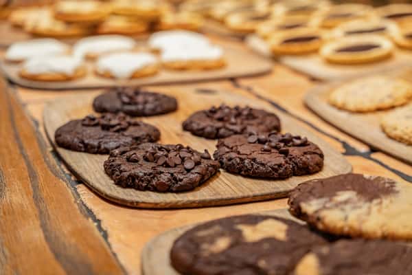 Assortment of freshly baked cookies displayed on wooden trays, featuring rich double chocolate chip cookies in the foreground, surrounded by frosted, jam-filled, and classic varieties on a rustic wooden table—ideal for a bakery or dessert café setting.