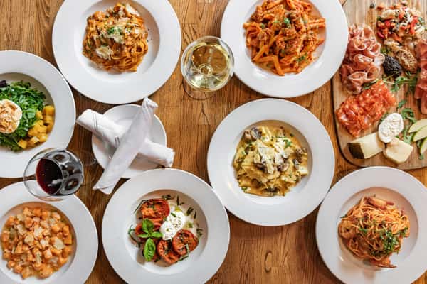Top-down view of a full Italian table spread including pasta, salad, burrata, risotto, charcuterie, and wine.