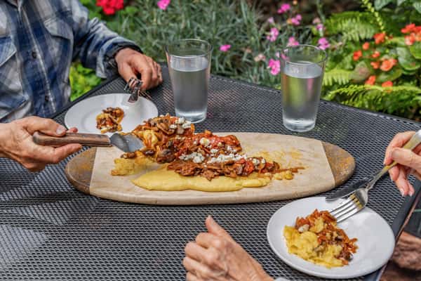 Outdoor dining scene with a group sharing a rustic polenta board topped with sautéed mushrooms, roasted tomatoes, and crumbled blue cheese, served al fresco with glasses of water and surrounded by blooming garden flowers.