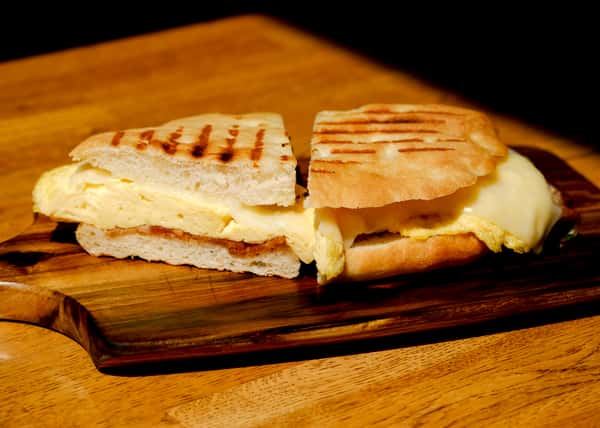 Grilled breakfast panini filled with fluffy eggs and melted cheese on ciabatta bread, served on a wooden board for a hearty café breakfast option.