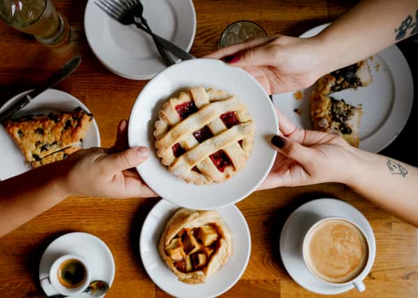 Hands presenting a small lattice-topped berry tart surrounded by plates of assorted pastries, espresso, and a latte on a wooden café table.