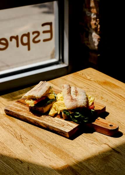 Sunny café window scene featuring a breakfast sandwich with scrambled eggs, spinach, and tomato on toasted ciabatta, served on a wooden board.